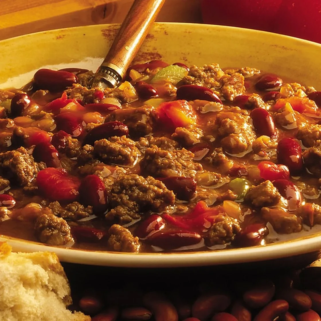 Bulk chili con carne with ground beef, red beans, and diced peppers in a yellow foodservice bowl with wooden spoon.