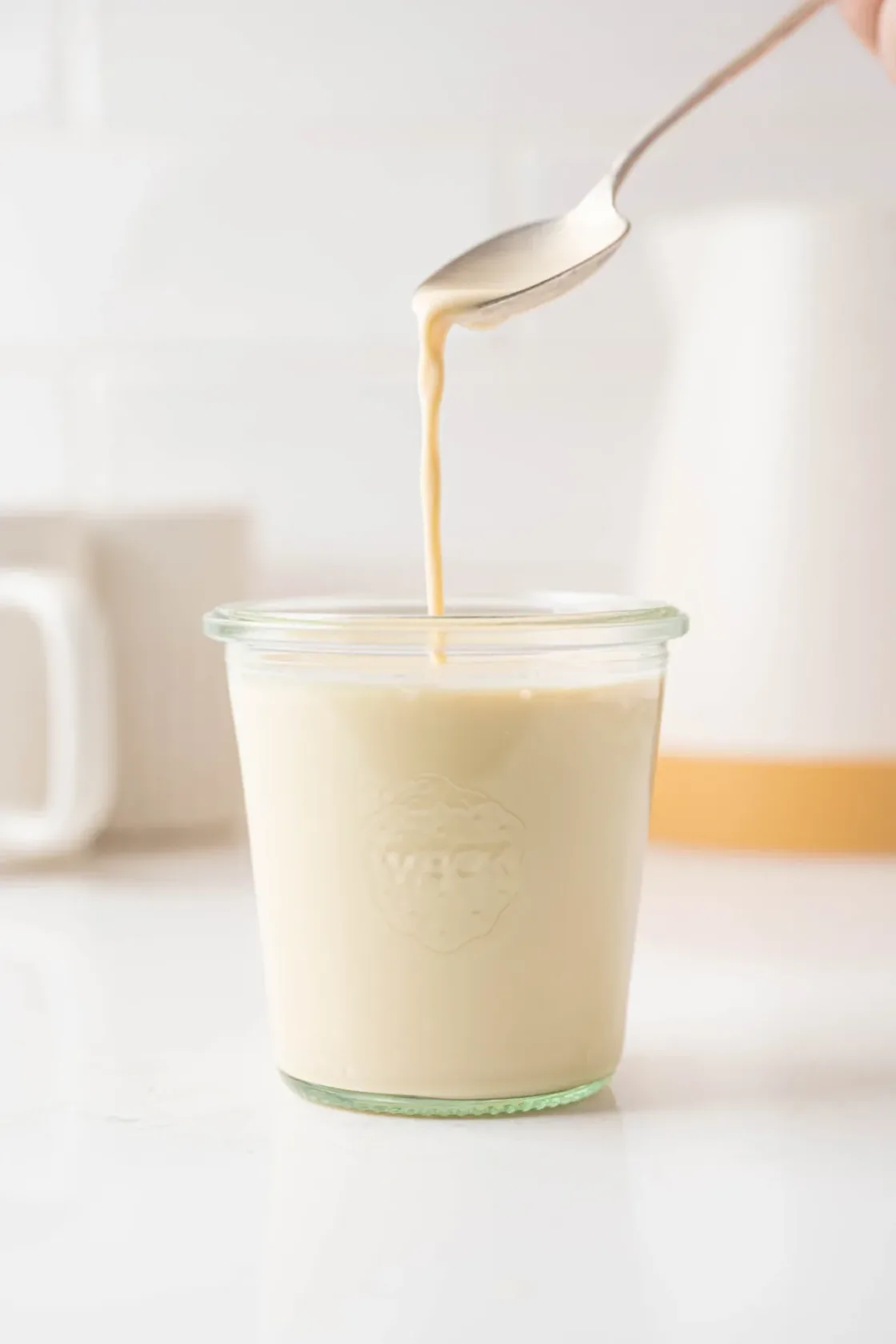 Golden evaporated milk being poured from a spoon into a glass jar, ideal for food service and commercial food preparation.