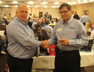 Two foodservice industry professionals shake hands at a wholesale food trade conference event with attendees and booths visible.