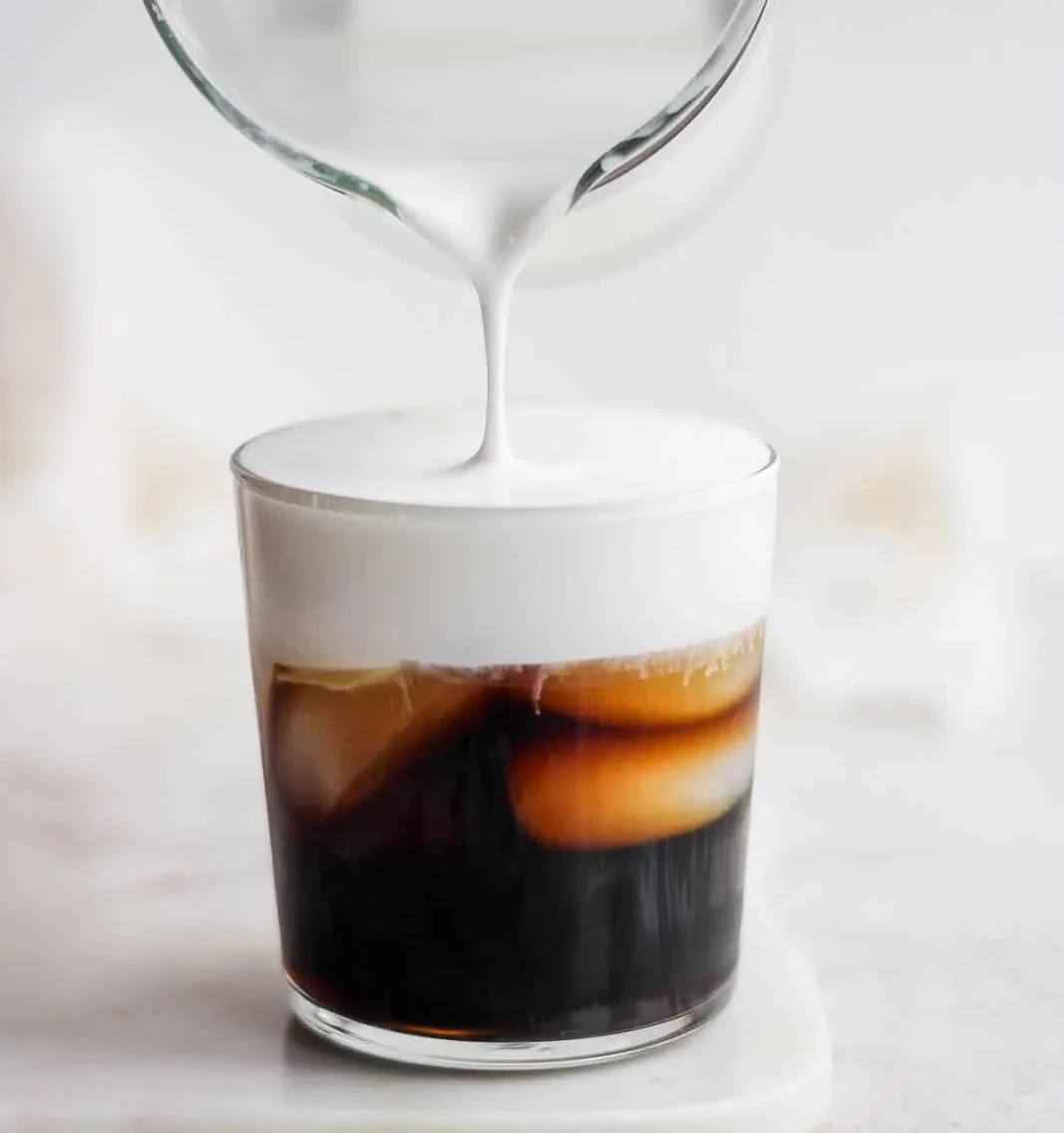 Cold foam milk being poured into dark iced coffee in a glass, professional beverage preparation for foodservice.